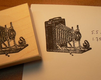 May include: A vintage-style camera with bellows and a lens, printed on a wooden block. The block is 1.75 x 2 inches.