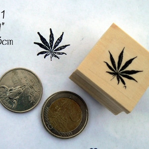 Könnte beinhalten: Ein Holz-Stempel mit einem schwarzen Marihuana-Blatt-Design. Der Stempel misst 1x1 Zoll oder 3x3 cm. Der Stempel wird neben zwei Münzen zur Größenvergleich gezeigt.