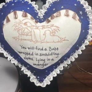 May include: A blue heart-shaped pillow with white lace trim and a white fabric center. The center features an embroidered image of the nativity scene with the text "You will find a Babe wrapped in swaddling cloths, Lying in a manger".