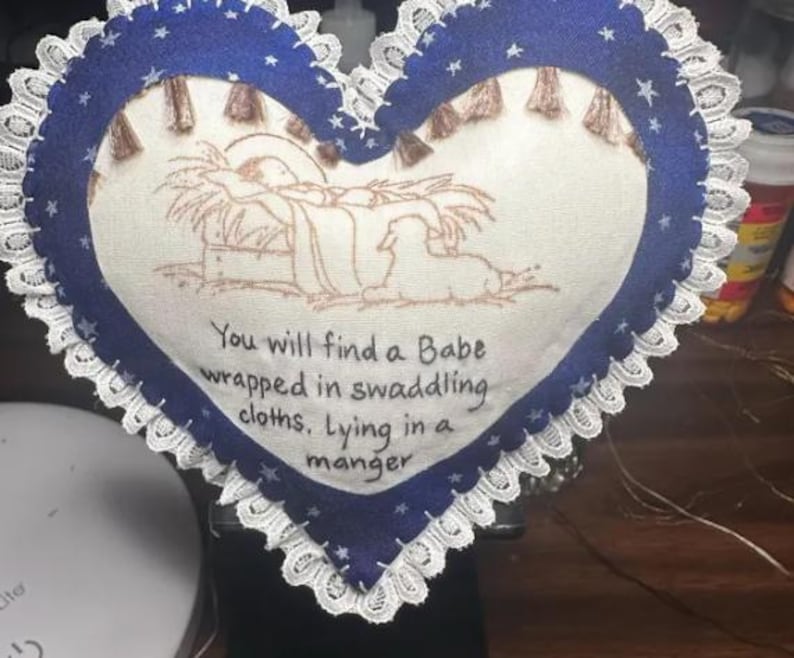 May include: A blue heart-shaped pillow with white lace trim and a white fabric center. The center features an embroidered image of the nativity scene with the text "You will find a Babe wrapped in swaddling cloths, Lying in a manger".