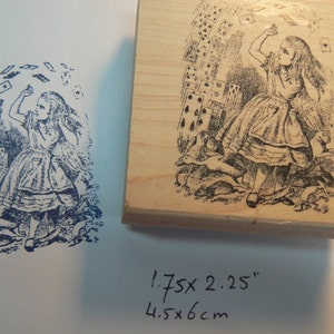 Könnte beinhalten: Ein Holz-Stempel mit einer Schwarzweiß-Illustration von Alice aus Alice im Wunderland. Alice steht mit erhobenen Armen, umgeben von Spielkarten. Der Stempel misst 4,5 x 6 cm.