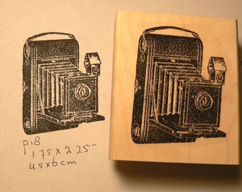 May include: A vintage-style wooden stamp featuring a black and white illustration of a folding camera. The stamp measures 1.75 x 2.25 inches or 4.5 x 6 cm.