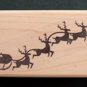 May include: A wooden stamp with a silhouette of a sleigh pulled by four reindeer. The reindeer are in a line, with the sleigh at the front. The stamp is a light brown color and has a smooth surface.