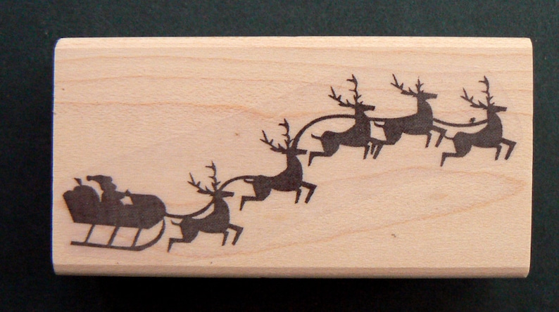 May include: A wooden stamp with a silhouette of a sleigh pulled by four reindeer. The reindeer are in a line, with the sleigh at the front. The stamp is a light brown color and has a smooth surface.