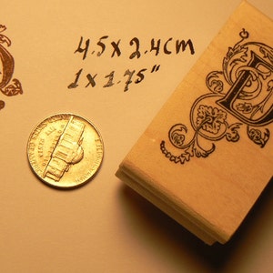 May include: A wooden rubber stamp with a black ink design of a letter D with decorative flourishes. The stamp measures 1 x 1.75 inches and 4.5 x 2.4 cm.