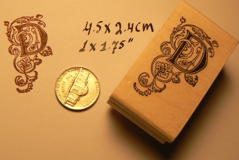 May include: A wooden rubber stamp with a black ink design of a letter D with decorative flourishes. The stamp measures 1 x 1.75 inches and 4.5 x 2.4 cm.