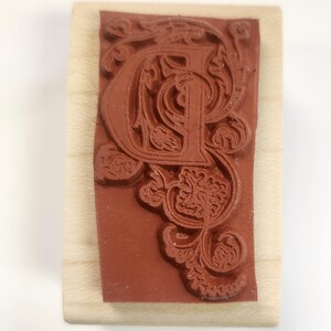 May include: A wooden mounted rubber stamp with a decorative, ornate, and intricate design of the letter "P" in a brown color.