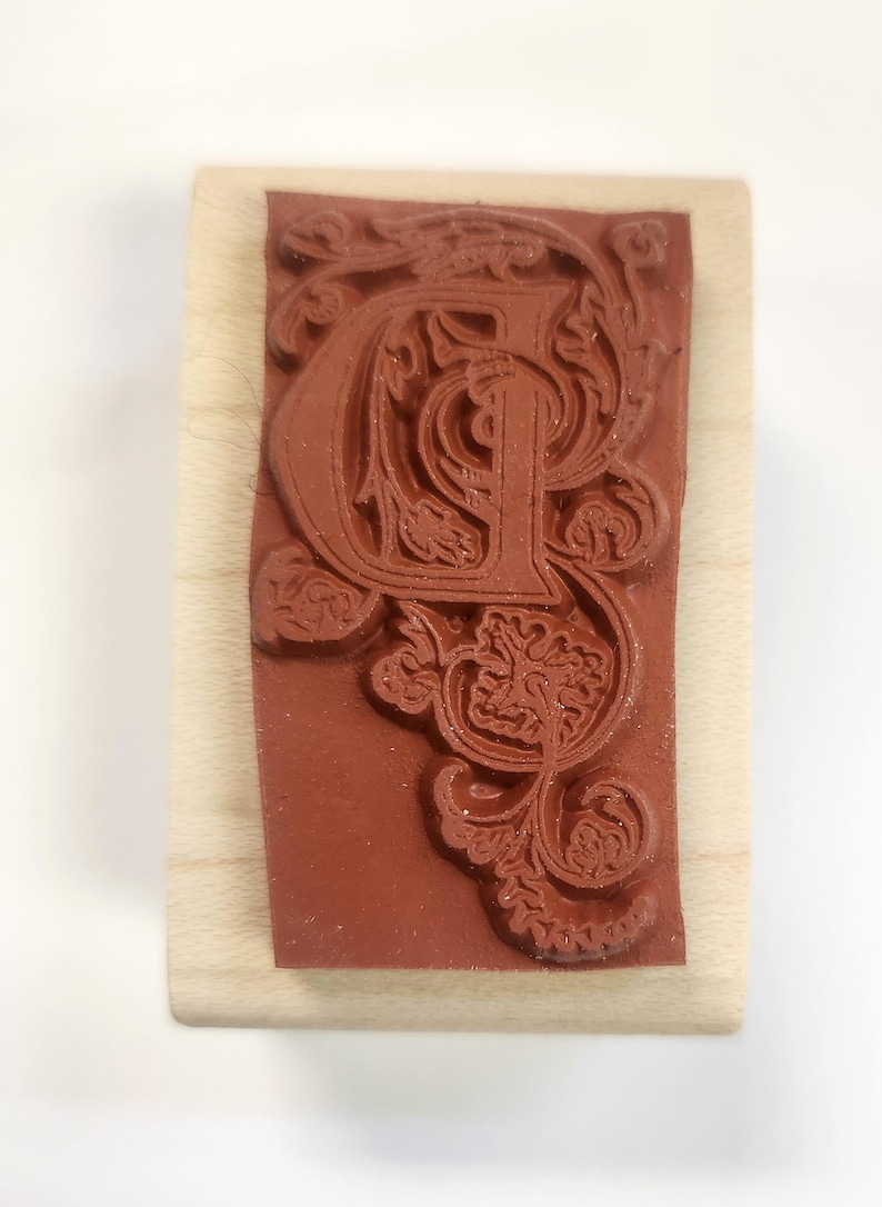 May include: A wooden mounted rubber stamp with a decorative, ornate, and intricate design of the letter "P" in a brown color.