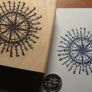 May include: A wooden stamp with a black ink compass design. The compass has a circular design with a star in the center and 32 points around the edge. The points are labeled with words in a circular pattern. The stamp is 2 inches in diameter.