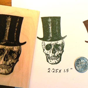 May include: A wooden stamp with a black skull wearing a top hat. Two skull prints in green and brown ink are shown, along with a quarter for size reference. The stamp is 2.25 x 1.5 inches.