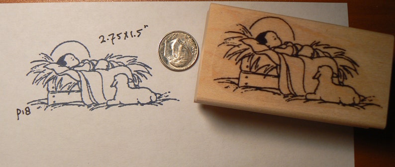 May include: A wooden rubber stamp with a black and white illustration of a baby in a manger with two sheep. The stamp measures 2.75 x 1.5 inches.