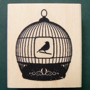 Puede incluir: Un sello de goma negro y blanco con un dise&ntilde;o de p&aacute;jaro en una jaula. La jaula es redonda y tiene una base decorativa. El p&aacute;jaro est&aacute; posado en una rama dentro de la jaula.