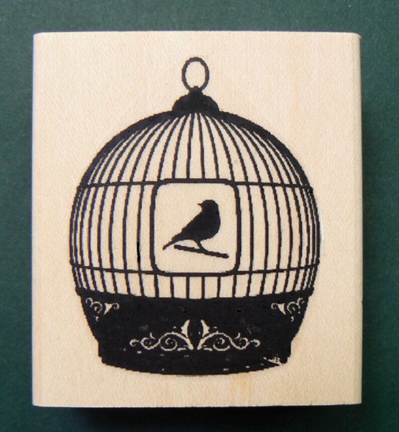 Puede incluir: Un sello de goma negro y blanco con un dise&ntilde;o de p&aacute;jaro en una jaula. La jaula es redonda y tiene una base decorativa. El p&aacute;jaro est&aacute; posado en una rama dentro de la jaula.