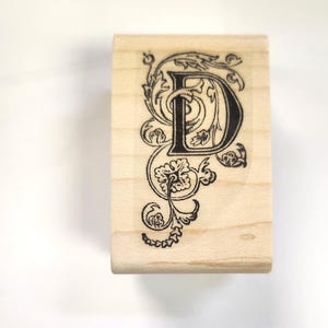 May include: A wooden stamp with a black and white design of the letter "D" with decorative flourishes.