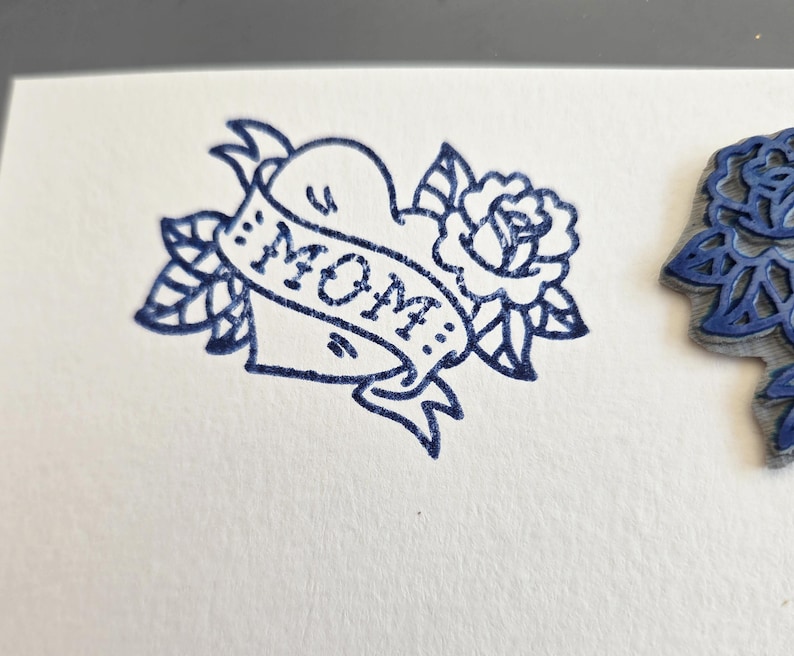 May include: A blue ink stamp of a heart with a banner that reads "MOM" surrounded by leaves and a rose. The stamp is on white paper. The image is a close-up of the stamp.