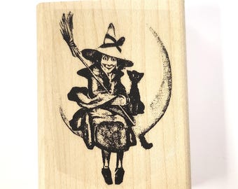 Könnte beinhalten: Ein Holz-Stempel mit einem Schwarzweißbild einer Hexe, die auf einem zunehmenden Mond mit einer Katze reitet. Die Hexe hält einen Besen und trägt einen spitzen Hut.