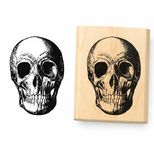 May include: A rectangular wooden stamp featuring a detailed black ink illustration of a human skull. Beside the stamp is a matching black ink impression of the same skull design, showing the anatomical features of the cranium and teeth.