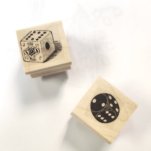 Könnte beinhalten: Zwei Holzstempel mit schwarzen Tintenmotiven. Ein Stempel zeigt einen Würfel mit einem Pik-Buben auf einer Seite und Punkten auf der anderen. Der andere Stempel zeigt einen Würfel mit Punkten auf allen Seiten.