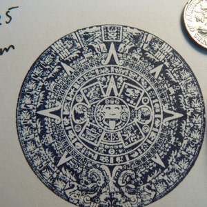 May include: A blue and white circular stamp with an intricate design, featuring a central sun with a face and a ring of glyphs around it. The stamp is 2 inches in diameter and is labeled "P25".