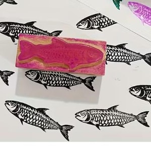 May include: A pink fish-shaped stamp with a wooden base is surrounded by black and white fish prints. A purple fish print is also visible. The fish prints are detailed with scales and fins, creating a nautical theme.