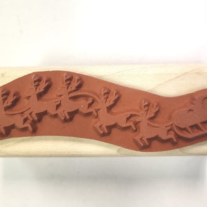May include: A wooden mounted rubber stamp with a design of a sleigh pulled by reindeer. The stamp is brown and the design is red.