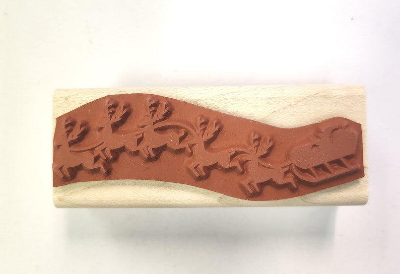 May include: A wooden mounted rubber stamp with a design of a sleigh pulled by reindeer. The stamp is brown and the design is red.