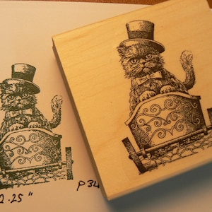 Può includere: Un timbro in gomma di legno con un'illustrazione in bianco e nero di un gatto che indossa un cappello a cilindro e un papillon, seduto su una carrozza con un design decorativo. Il timbro misura 2 x 2,25 pollici.