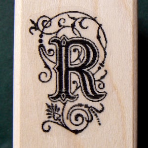 May include: A wooden stamp with a black ink design of the letter "R" surrounded by decorative flourishes.