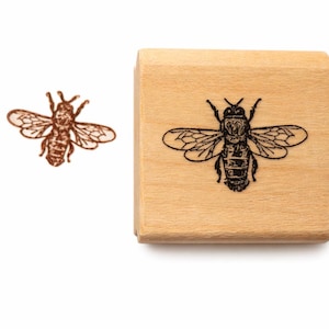 May include: A rectangular wooden rubber stamp featuring a detailed black ink illustration of a honeybee on its surface. To the left of the stamp, a brown ink impression of the same honeybee design is displayed, showing the intricate wing and body details.