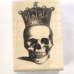 May include: A wooden stamp with a black and white image of a skull wearing a crown. The skull is detailed with a prominent jawline and teeth.