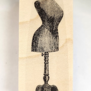 May include: A vintage-style wooden stamp with a black and white illustration of a dress form on a stand.