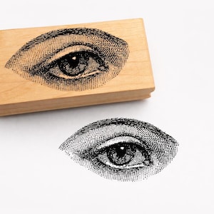 Op de afbeelding: Een houten stempel met een gedetailleerd zwart-wit oogontwerp. De stempel is rechthoekig en ligt naast een afdruk van het oog. Het oogontwerp is in een vintage illustratiestijl.