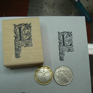 Könnte beinhalten: Ein Stempel aus Holz mit einem schwarz-weißen Design des Buchstabens L, umgeben von verzierten Schnörkeln. Der Stempel befindet sich auf einer weißen Oberfläche mit zwei Münzen daneben.