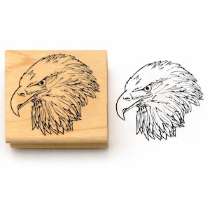 May include: A square wooden rubber stamp featuring a detailed black line art illustration of a bald eagle head in profile. The design shows the eagle facing left with sharp, defined feathers and a hooked beak, presented on a light wood block.
