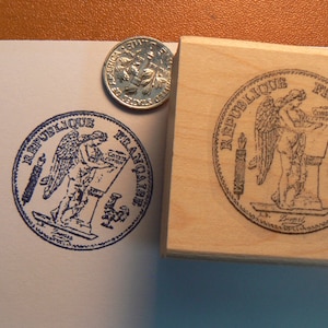 May include: A wooden stamp with a blue ink design of a winged figure holding a scroll with the text "REPUBLIQUE FRANCAISE" around the edge. The stamp is 1.25 inches or 3.5 cm in diameter.