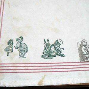 May include: White tea towel with red stripes and green and black printed illustrations of Alice in Wonderland characters, including the Dodo, the Mad Hatter, and the March Hare.
