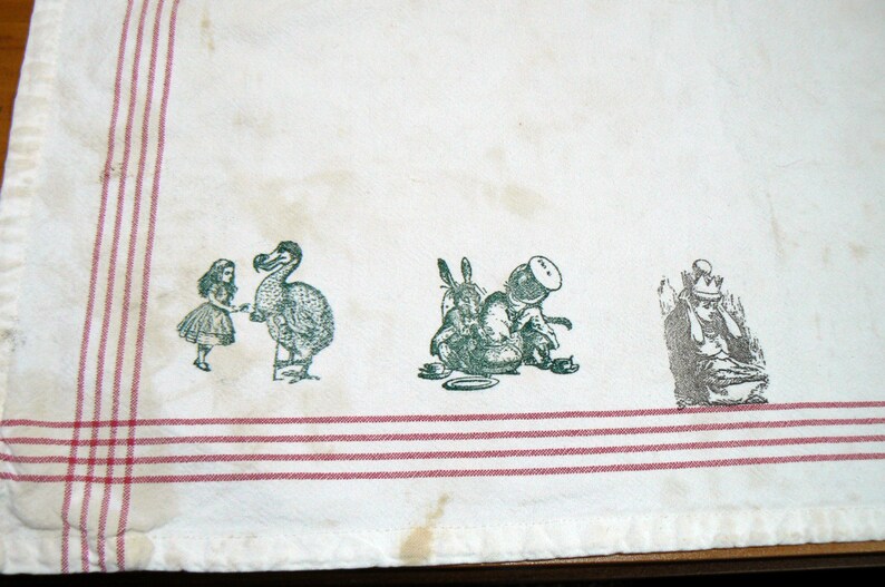 May include: White tea towel with red stripes and green and black printed illustrations of Alice in Wonderland characters, including the Dodo, the Mad Hatter, and the March Hare.