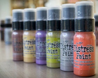 Puede incluir: Una fila de diez botellas pequeñas de pintura Distress en varios colores, incluyendo marrón, amarillo, morado, verde, gris y naranja. Las botellas están etiquetadas con el nombre de la marca y el color de la pintura.