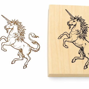 May include: A rectangular wooden rubber stamp featuring a detailed black ink illustration of a rearing unicorn with a flowing mane and tail. Beside the stamp is a separate brown ink impression of the same unicorn design on a plain white background.