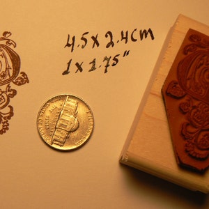 May include: A wooden stamp with a brown, intricate design of a letter "D" with floral accents. The stamp is 4.5 x 2.4 cm or 1 x 1.75 inches. A US penny is included for scale.