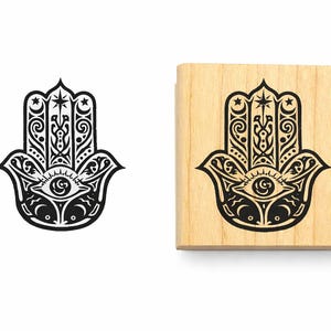 Pu&ograve; includere: Un timbro quadrato in legno con un design a mano di Hamsa in inchiostro nero, caratterizzato da filigrana complessa, un occhio centrale e motivi celesti con stelle e lune. Accanto al timbro si trova un'impronta nera del medesimo disegno.