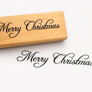 Op de afbeelding: Een houten stempel met de woorden "Merry Christmas" in elegant schrift. De stempel is lichtbruin en de tekst is zwart. Een afdruk van de tekst staat eronder op een witte ondergrond.
