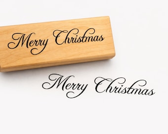 May include: A wooden stamp with the words "Merry Christmas" in elegant script. The stamp is light brown, and the text is black. A stamped impression of the text is below the stamp on a white surface.