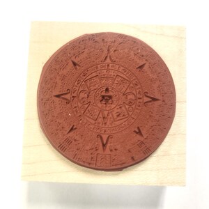 May include: A wooden block with a round, red rubber stamp of the Aztec calendar. The stamp is detailed with intricate carvings and glyphs.