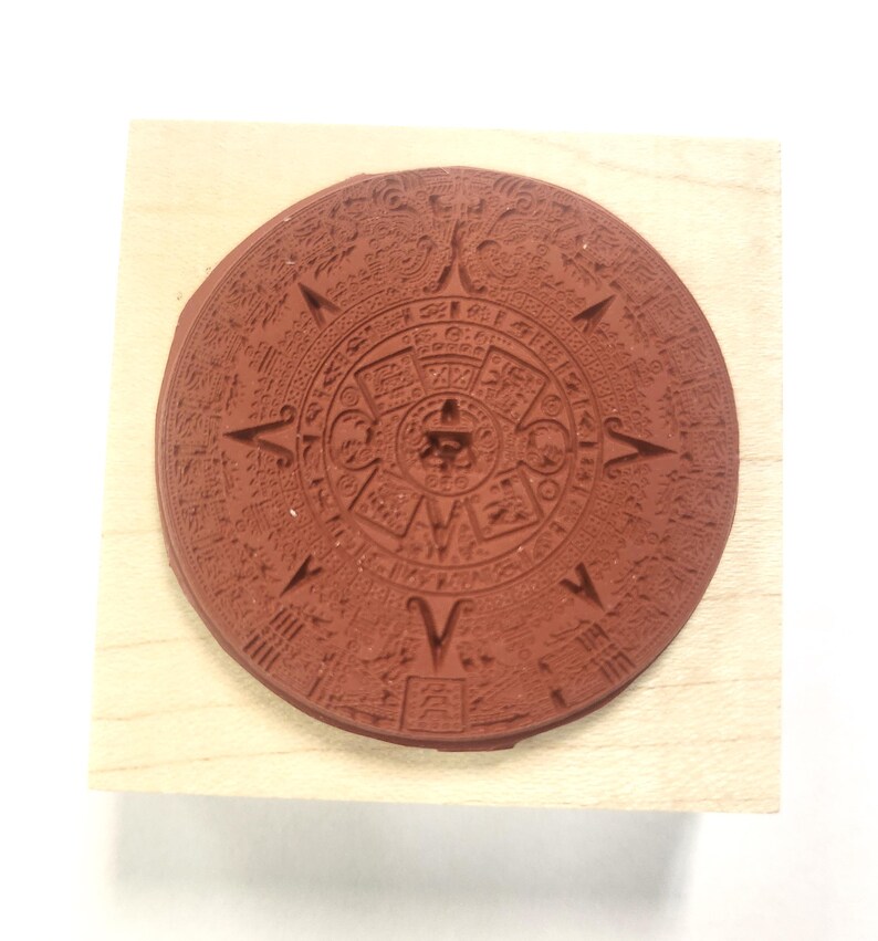 May include: A wooden block with a round, red rubber stamp of the Aztec calendar. The stamp is detailed with intricate carvings and glyphs.