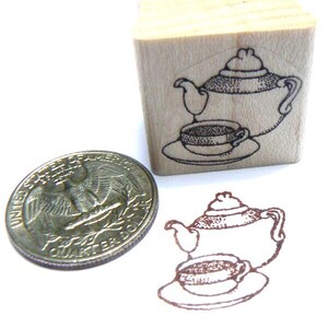 May include: A wooden rubber stamp with a black and white illustration of a teapot and a teacup on a saucer. The stamp is about the size of a US quarter.
