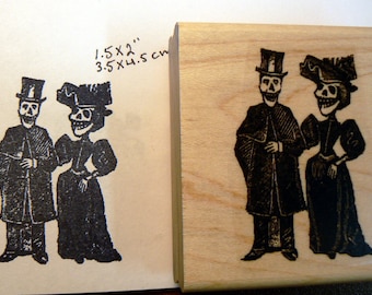 May include: A wooden stamp with a black and white image of two skeletons dressed in formal attire. The skeletons are wearing top hats and have a skull and crossbones design. The stamp measures 1.5 x 2 inches and 3.5 x 4.5 cm.