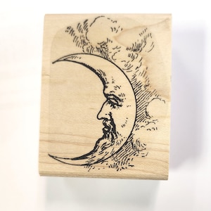 May include: A wooden stamp featuring a black and white illustration of a crescent moon with a face. The moon is surrounded by clouds.