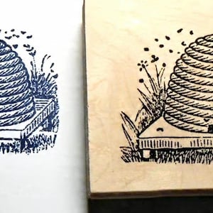 May include: A square wooden rubber stamp featuring a detailed black ink illustration of a domed beehive resting on a platform, surrounded by stylized grass and flying bees, shown alongside a crisp blue ink impression of the same design.