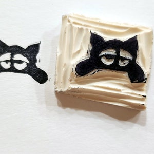 Puede incluir: Un sello de goma negro con una imagen tallada de un gato con ojos grandes. El gato está mirando hacia abajo y tiene una expresión triste.